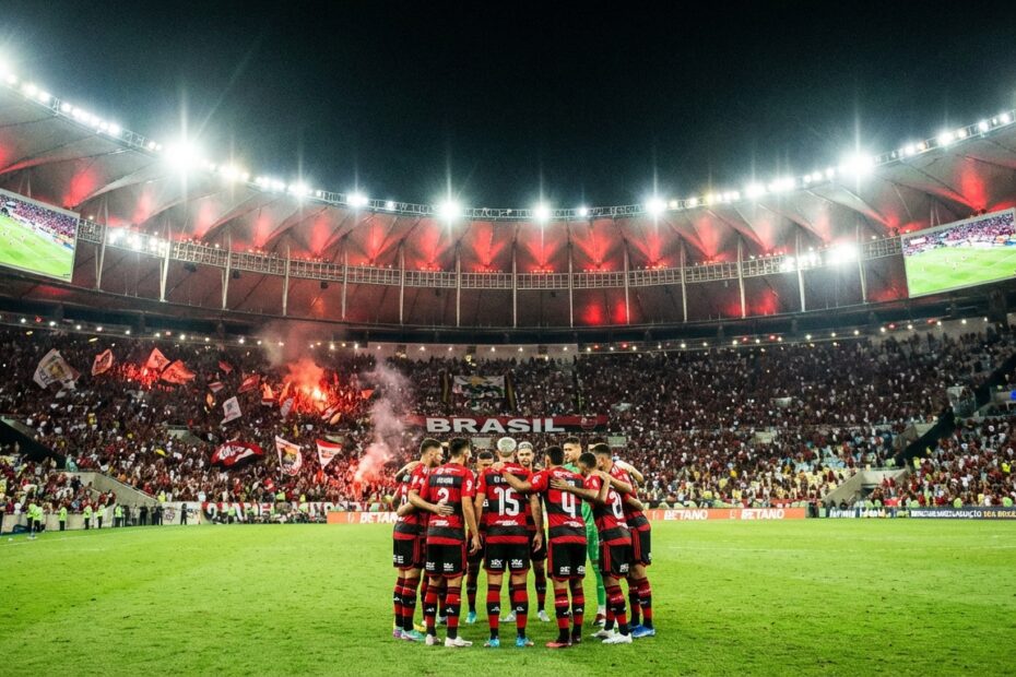 Jogadores com a camisa vermelha e preta em campo iluminado de estádio