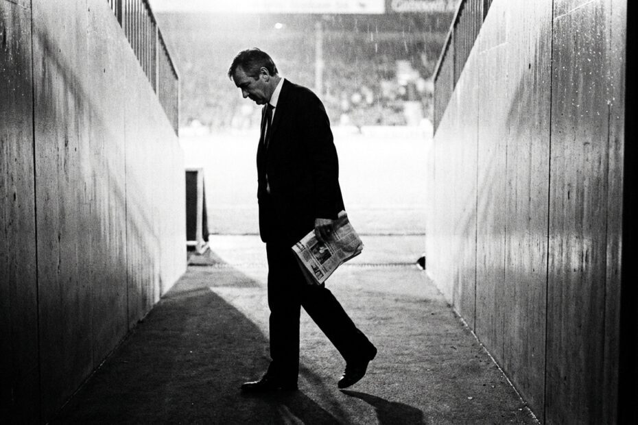 Executivo de futebol caminhando sozinho em túnel de estádio com expressão preocupada